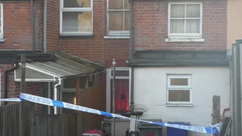 Jamie Niblock/BBC A terraced house is in the middle and the first floor is painted white and the top floor is made of red bricks. There is a blue and white police tape outside of the building. 
