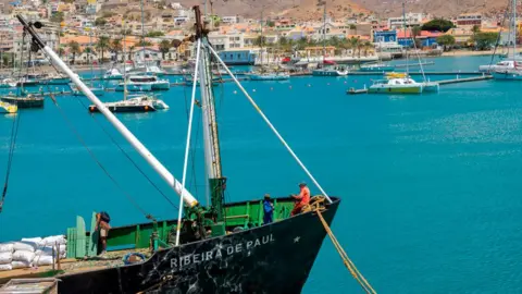 Getty Images Para pekerja memuat barang ke kapal di pelabuhan Mindelo di Tanjung Verde. Kapal pesiar dapat dilihat di latar belakang. Laut aquamarine tenang. 