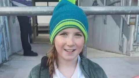 Rathdrum Rugby Club A young girl sits and smiles at the camera holding a white, green and black rugby ball. She is wearing a green and blue beanie hat, a green coat and rugby shirt