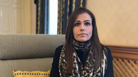 Lucy Domaille - A woman with long brown hair, looking towards the camera. She is wearing a leopard print scarf over a navy blue blouse. 