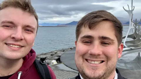 Elliot Griffiths and Zak Nelson facing the camera and smiling in a selfie in front an abstract metal sculpture with water and mountains in the background