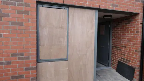 A close up of the entrance to a house with its windows boarded up in north Belfast following sectarian attacks on 21 May.   The front door of the property is open and the building is made of new red brick. 