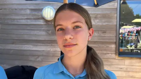 A girl with brown hair tied in a pony tail wearing a blue polo school uniform looking at the camera. Behind is a wooden building with a window on the right and a outdoor light on the right and a dark blue umbrella above.