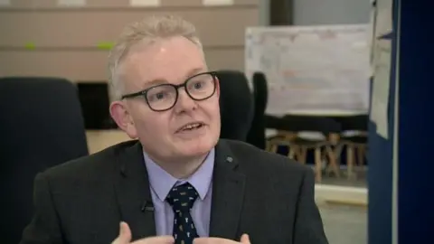 Ian McCord with short white hair wearing black glasses, a dark jacket, pink shirt and black tie. He is in a large hall and is talking.