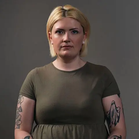 BBC Scotland Robyn, who has a blonde bob, is staring straight at the camera. She is wearing a khaki green dress and has tattoo on her arms. 