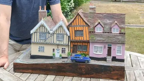 A model of a row of cottages. There are two cottages, one pink and one cream, with a smaller crooked cottage sitting in between them in an orange colour. There is a blue car parked outside the orange home. A man's arms can be seen resting on the table which the model sits on.