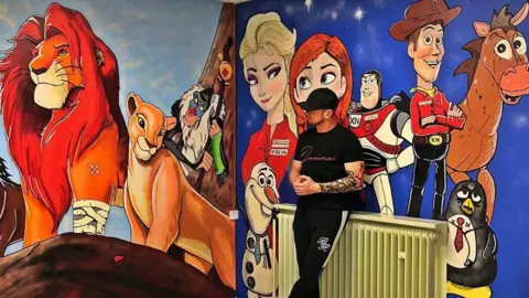 Adam Salisbury Artist Adam Salisbury wearing a black baseball cap, black t-shirt and black and white joggers, stands in the middle of a mural of Toy Story and the Lion King characters on two walls in the children's ward at Blackpool Victoria Hospital.