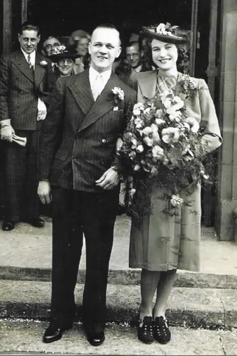 Drew family photograph Stan and Beatrice pose in a black and white photo. Stan is wearing a pinstripe suit with a button hole, Beatrice holds a large bouquet of orchids and is wearing a period dress and jacket with dark hat and shoes