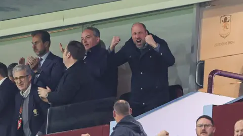 Getty Images Prince William is standing with his fists in the air in celebration, with his face mid-cheer. There is a group of men to his right that all appear to be clapping