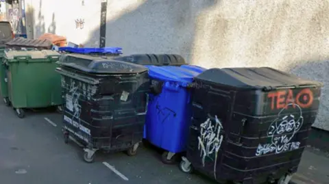 LDRS A collection of industrial waste and recycling bins covered in graffitti next to a pavement. 