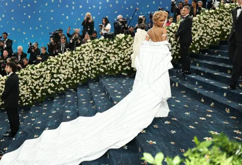 MICHAEL LOCCISANO/GETTY IMAGES A wide shot of Tyla on the blue carpet, who is wearing a flowy white dress with a long tail  and sporting a blonde-pixie style cut.