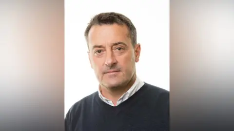 St Albans City and District Council A studio-style head-and-shoulders portrait shows Edgar Hill positioned against a plain white background. He is wearing a dark crew-neck sweater layered over a white shirt.