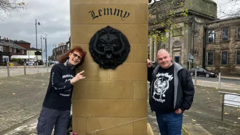A couple in Motorhead themed T-shirts stand either side of the plinth of the Lemmy statue, which has Lemmy engraved on it. Also on the plinth is is Motorhead's large black motif, which is known as the Snaggletooth or the War Pig and sometimes the Iron Boar. The statue is in a square, with roads and buildings surrounding it.