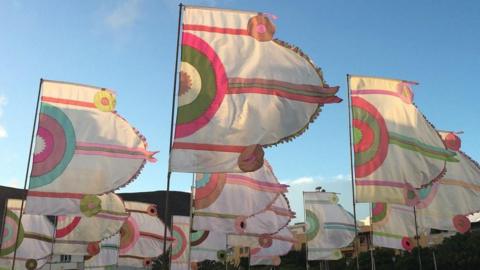 White flags with concentric half-circles and horizontal lines, in pink, green and brown, blowing in wind at festival site.