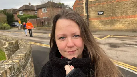 John Devine/BBC Francesca Pryar is looking at the camera. She has long dark hair over her shoulders. She is wearing a black fleece top and behind her are two people crossing the road with a house visible too.