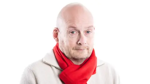 Midlands Air Ambulance Steve is wearing a cream jumper and red scarf and had a white background behind him. He has a bald head and a beard.