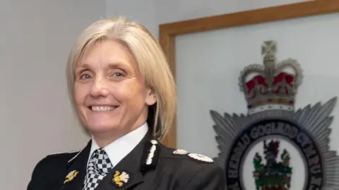 Amanda Blakeman in police uniform smiling at the camera. She has blonde bobbed hair and stands in front of framed North Wales Police crest