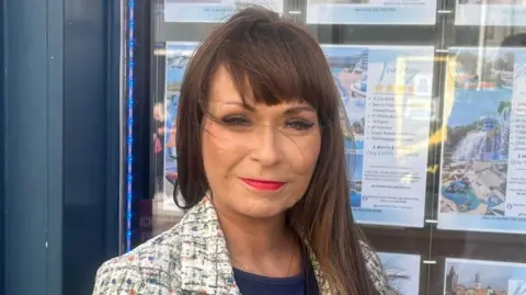 Jenny Elliott has long brown hair and is wearing a white multi-coloured jacket.
She is standing in front of her travel agency shop. 
