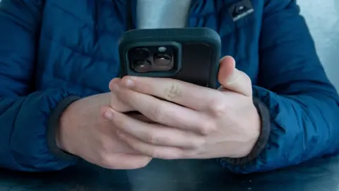 The torso of a male, dressed in a blue coat, observing a mobile phone in his hands