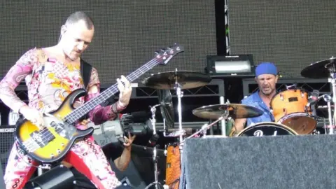 Jim Connolly/BBC Members of Red Hot Chili Peppers perform at Portman Road stadium. One member plays a bass guitar while another performs on the drums behind him.
