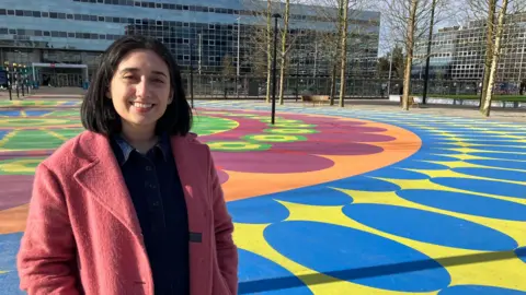 Amy Holmes/BBC A woman in a pink coat stands on a brightly painted outdoor area with bold blue, yellow, green, and orange patterns, with a modern glass‑fronted building and leafless trees in the background.