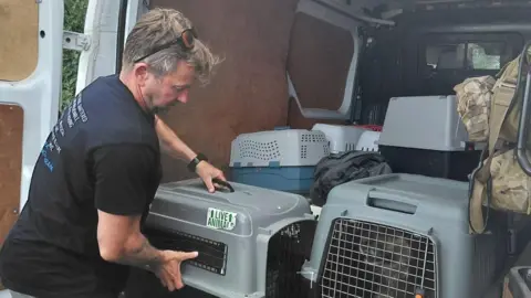 Nowzad Pen Farthing loading a grey crate containing an animal into a white van full of other crates. He is wearing a black T-shirt and has sunglasses on top of his head.