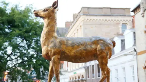 Prop-Search A golden statue of a hind deer, outside a building, showing buildings behind it, and a green tree to the left. 