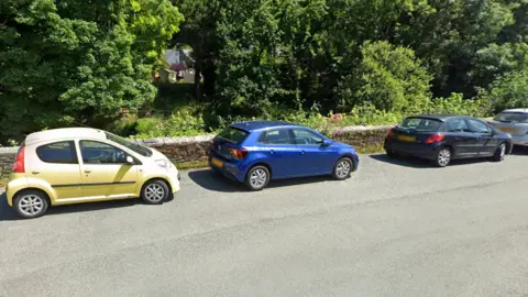 A street view of Market Road in Tavistock. There is a river wall next to which are parked several cars, and trees behind the wall