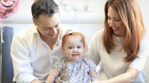 Team Margot A young girl with a tube glued to her cheek, which she holds up to her nose, sits next to her mother and father, who smile at her