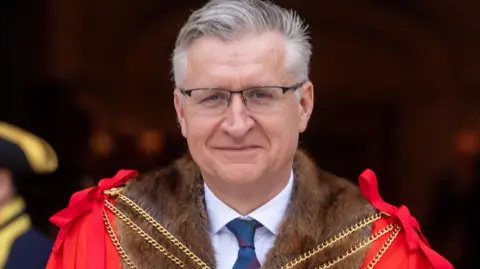 Ray Tang Media Alistair King wearing a red cloak lined with brown fur with gold chains over the top. He has grey hair and is wearing glasses