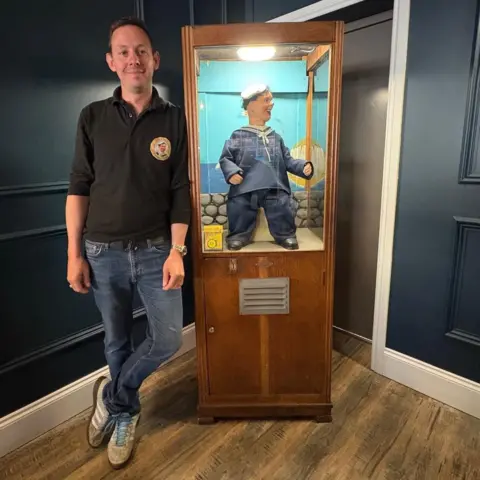 BBC / Kevin Shoesmith Gareth, a man in his 40s with short, brown hair, is standing next to a Laughing Sailor machine in a wooden cabinet.