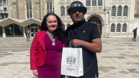 A woman in a purple dress and red jacket stands next to a man wearing a bowler hat with sunglasses on top as he hold a paper bag with the City of London emblem on it.