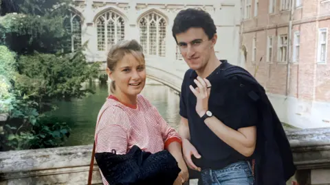 Two Rivers Media Rose Grant in red and white striped T shirt and Tim Burman in blue polo shirt and jeans, standing in front of a very old bridge somewhere in Cambridge. She is blonde, he has dark brown hair.
