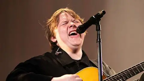 Getty Images Lewis Capaldi sings on a stage into a microphone as he plays a guitar. He has long brown hair and wears a black jacket. His eyes are shut as he performs. 