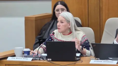 Kay Mason Billig is sitting behind a table in a council meeting. She has a laptop and a microphone in front of her. She is wearing a Christmas jumper, which is cream at the top, with red, blue and grey patterns lower down.