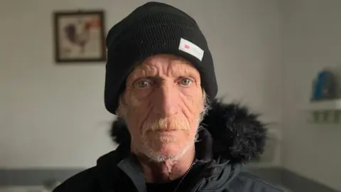 Ray is looking at the camera, he is an older man and is wearing a black woolen hat and a black coat. He has a short white beard. He is sitting inside in a room with white walls and a picture on the wall.
