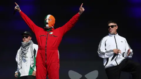 Kneecap members Mo Chara, DJ Próvaí and Móglaí Bap standing on a stage at Glastonbury. 