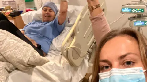 Lucy Goudswaard Joy lying in a hospital bed. She is dressed in a blue T-shirt and black trousers. She wears a blue hospital head covering. Her daughter Lucy is in the foreground, wearing a blue and white face mask. she has blonde hair and is looking into the camera. You can see medical equipment in the background.