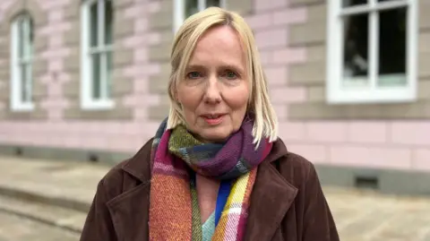 Catherine is wearing a dark coat and a multicoloured scarf stands outdoors in front of a light pink stone building with white-framed windows. The photo is taken at street level with shallow depth of field.