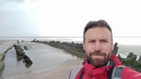 Dr Julian Whitewright Dr Julian Whitewright smiling at the camera (selfie style) in front of Cefn Sands shipwreck which is visible poking out of the sand, pools of water within the wreck show where sand has been hollowed out.