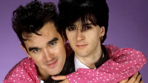 Getty Images Morrissey, avec ses cheveux noirs détachés et un T-shirt rose et blanc, serre dans ses bras Johnny Marr, qui a les cheveux noirs avec une frange au-dessus des yeux, et porte des boucles d'oreilles dorées et un T-shirt blanc sous une veste en cuir noire.