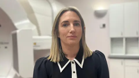 Dr Hanna has shoulder length blonde hair and is wearing a black top with white trim and standing in a hospital room, behind her the edge of a large white medical scanner can be seen.