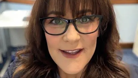 Close-up of a woman with long wavy brown hair wearing dark-framed glasses, indoors with a blurred background featuring a desk and chair.