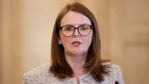 PA Media Caoimhe Archibald has shoulder-length red hair and large dark-framed glasses. She is wearing a beige jacket.