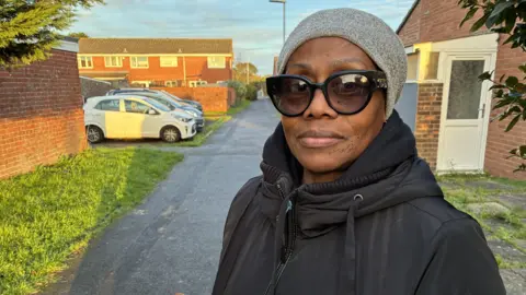 Image shows a woman with a grey beanie, black sunglasses, and a black coat.