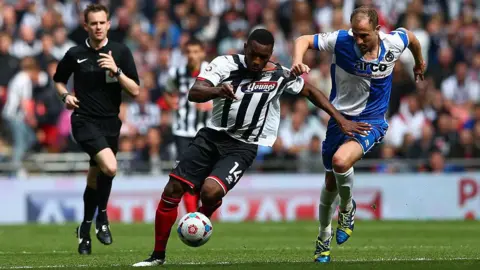 Getty Images Lenell John-Lewis in the centre of the picture, wearing a black and white striped Grimsby Town shirt and black shorts tussles for the ball with Mark McChrystal of Bristol Rovers, pictured on the right wearing a blue and white quadrant shirt and blue shorts. Just behind them and to the left of the image is the referee wearing an all-black kit.