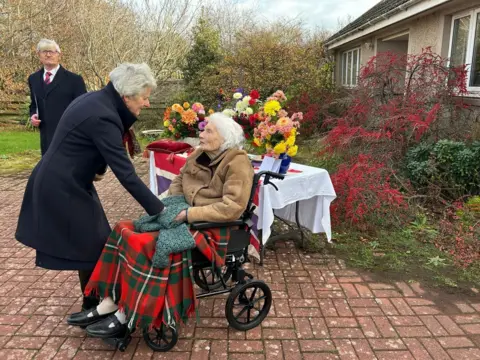 French Consulate A woman in a wheelchair with a tartan rug over her legs talks to another woman in a dark coat with grey hair