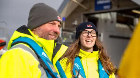 A man and a woman in RNLI-branded beanies. They are smiling and wearing a yellow hi-vis coat under a blue RNLI-branded lifejacket.