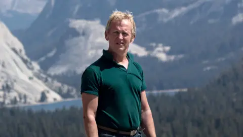Chesterfield Borough Council Stephen Lismore wearing a green polo shirt and jeans. He is standing in front of a backdrop of mountains and trees