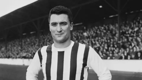 Getty Images A black-and-white image of British footballer Graham Williams pictured on a football pitch with stands full of fans in the background. He is wearing a striped football jersey.
